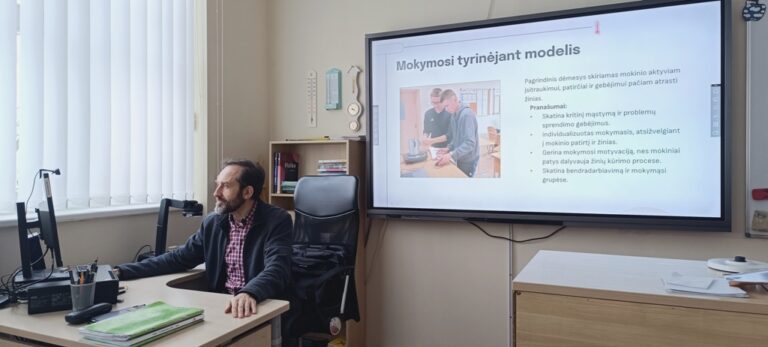 Mokytojas Vidas Jaunius sėdi prie stalo, ekrane matoma prezentacija apie mokymosi tyrinėjant modelį