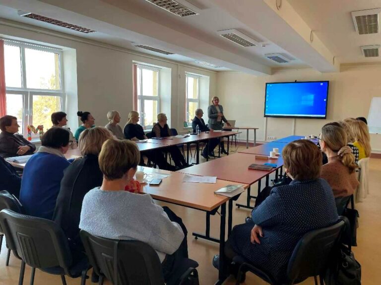 Apskritojo stalo diskusijos dalyviai sėdi prie stalų susirinkimų kambaryje, priekyje stovi pranešėja
